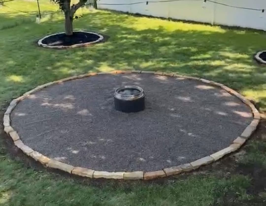 Circular gravel fire pit area surrounded by stone border in a green backyard.