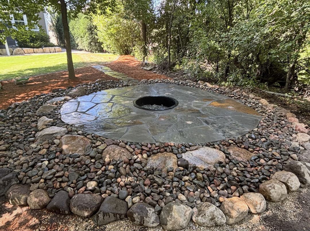Stone fire pit surrounded by a circular arrangement of pebbles and lush greenery.
