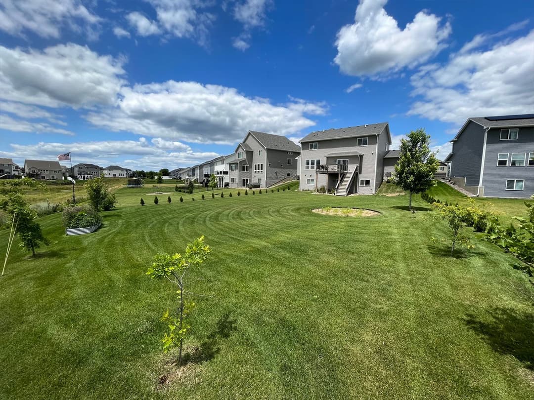 Lush green backyard with manicured lawn and modern homes under a blue sky with clouds.