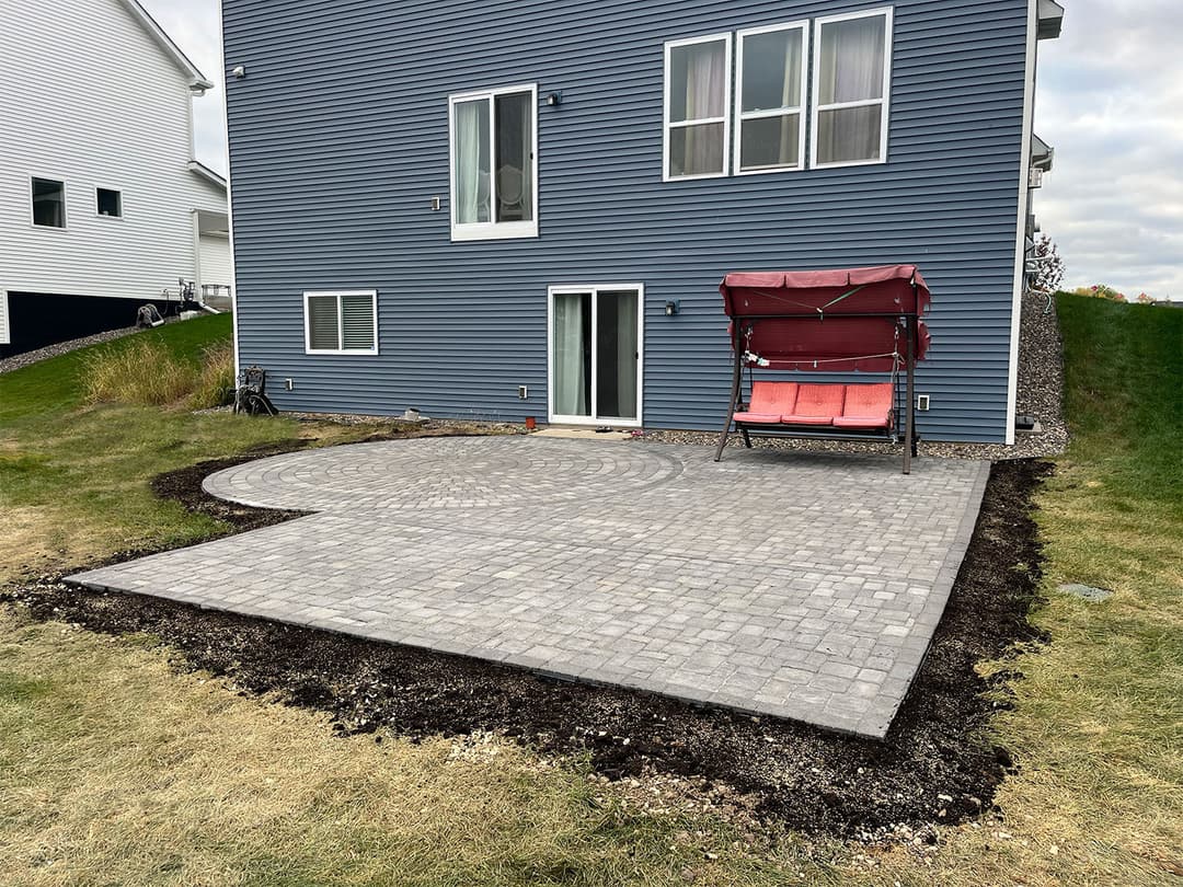 Paved patio area with a swing set beside a modern blue house in a residential neighborhood.