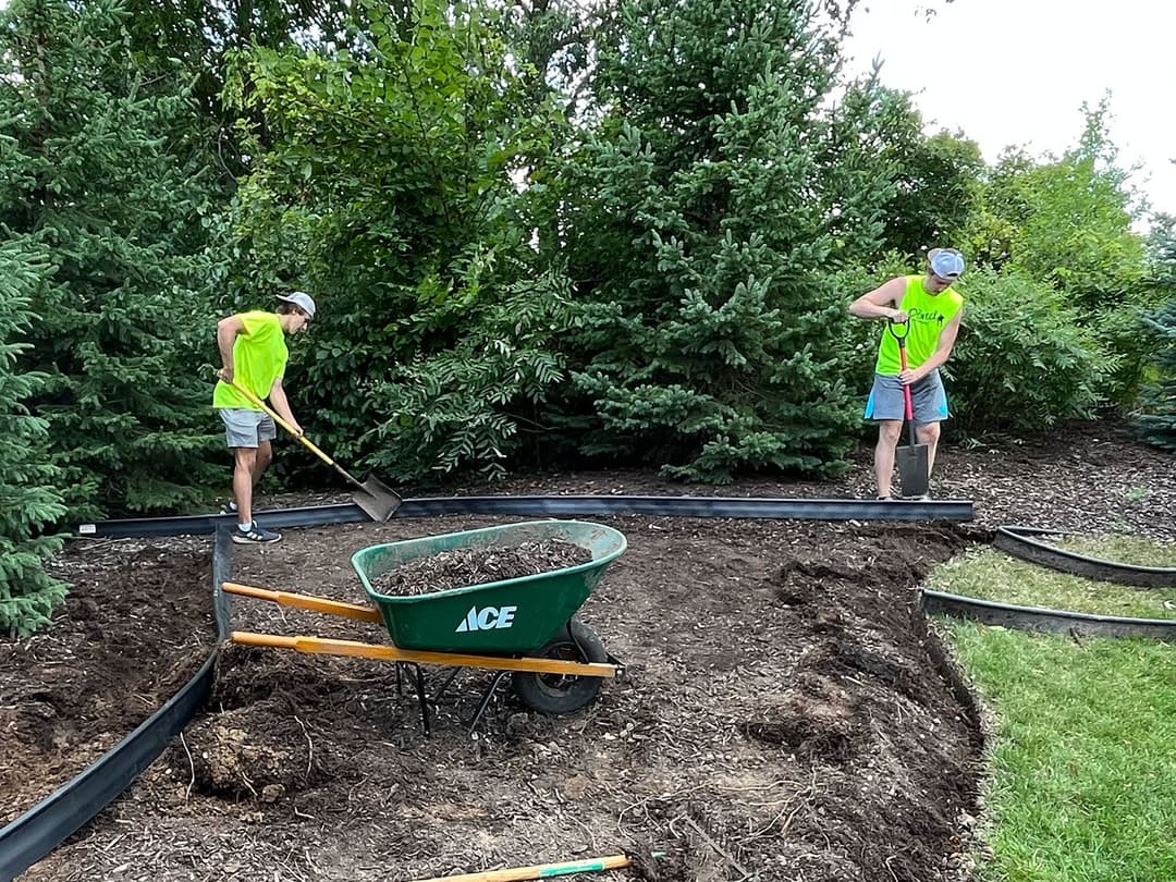 Two workers in bright vests landscaping a garden with a wheelbarrow and tools.