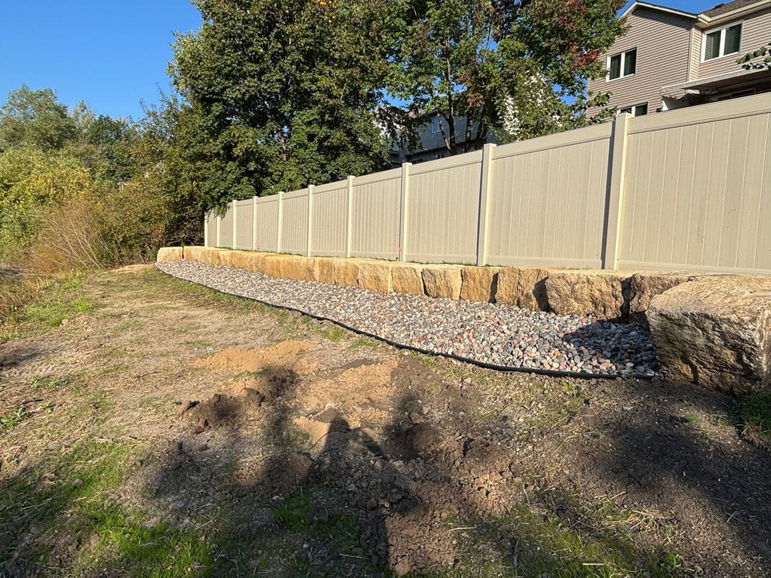 Landscape featuring a stone retaining wall with gravel along a fenced yard in autumn.