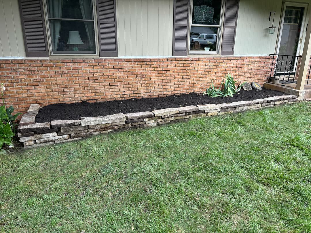 Stone retaining wall with fresh soil along a brick house foundation and lawn area.