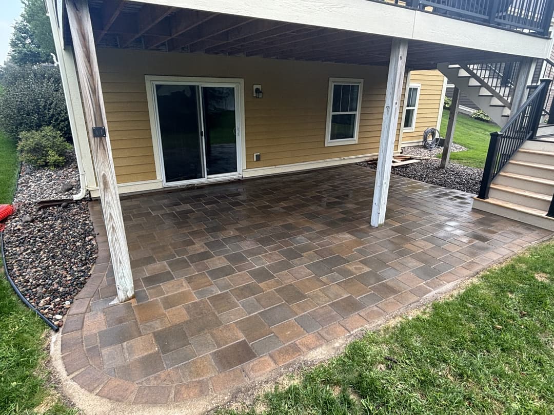 Paved patio under a raised deck, with grass and landscaping in the background.