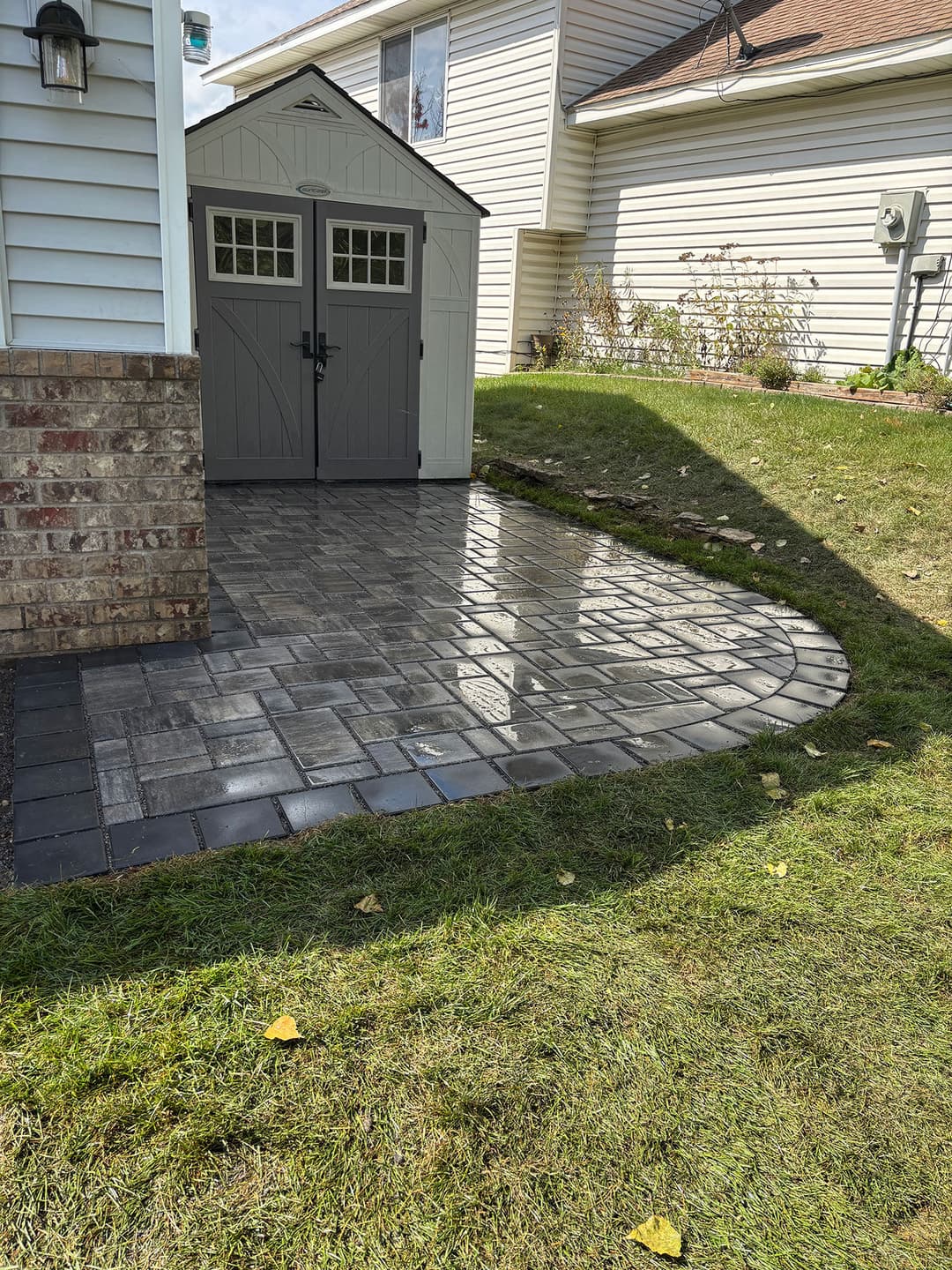 Paved patio with a shed, surrounded by grass and landscaping, under a clear sky.