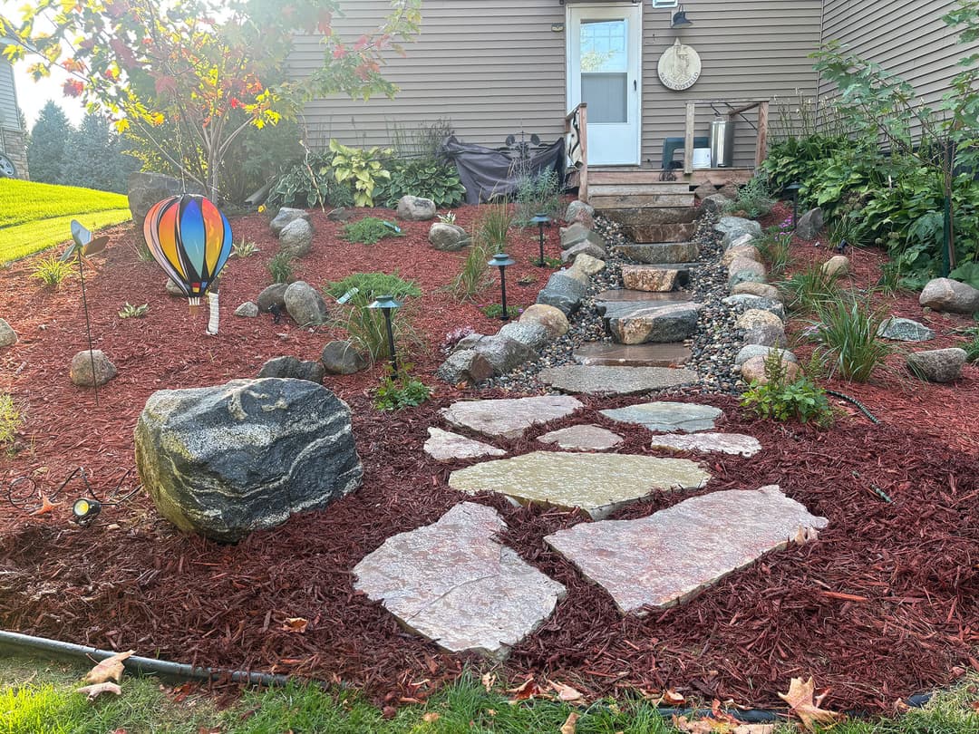 Colorful garden path with stone steps, rock features, and flowerbed near a house.