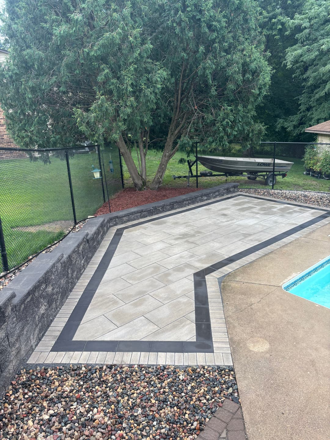 Rectangular patio with pavers and a black border beside a swimming pool and landscaped yard.