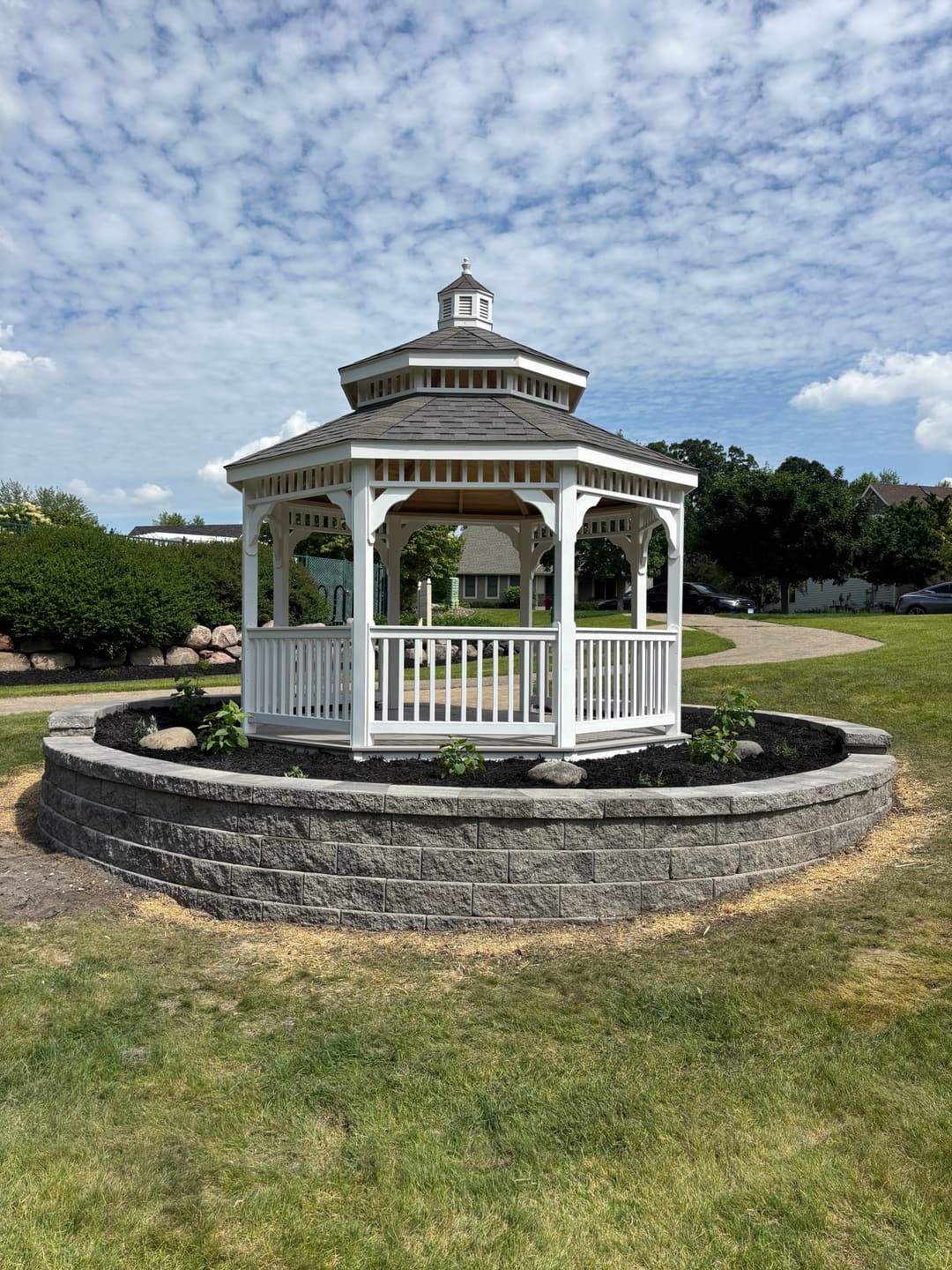 White gazebo surrounded by landscaped garden on a sunny day with blue sky and clouds.