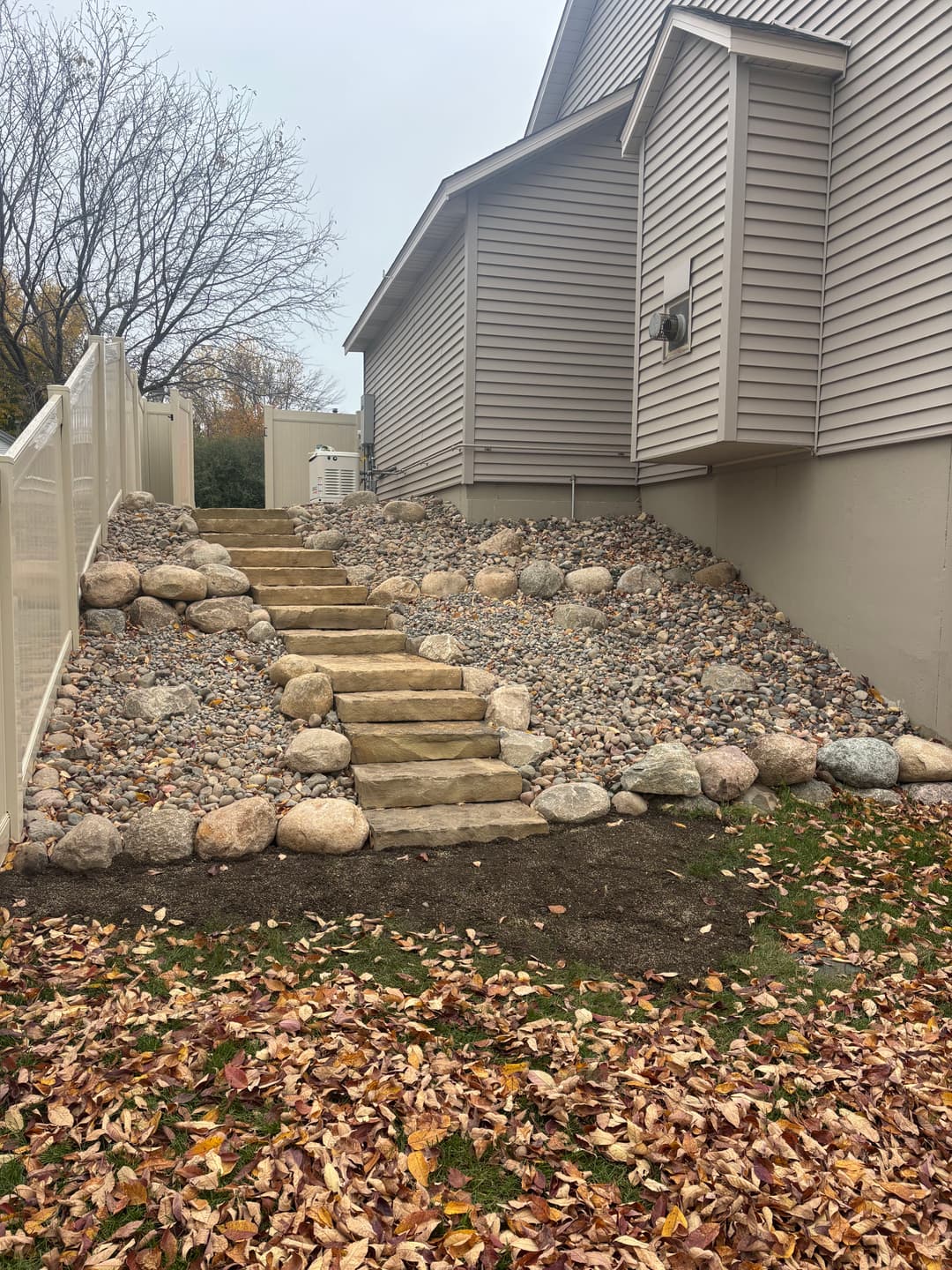 Stone pathway with steps leading up a landscaped hill beside a house, surrounded by autumn leaves.