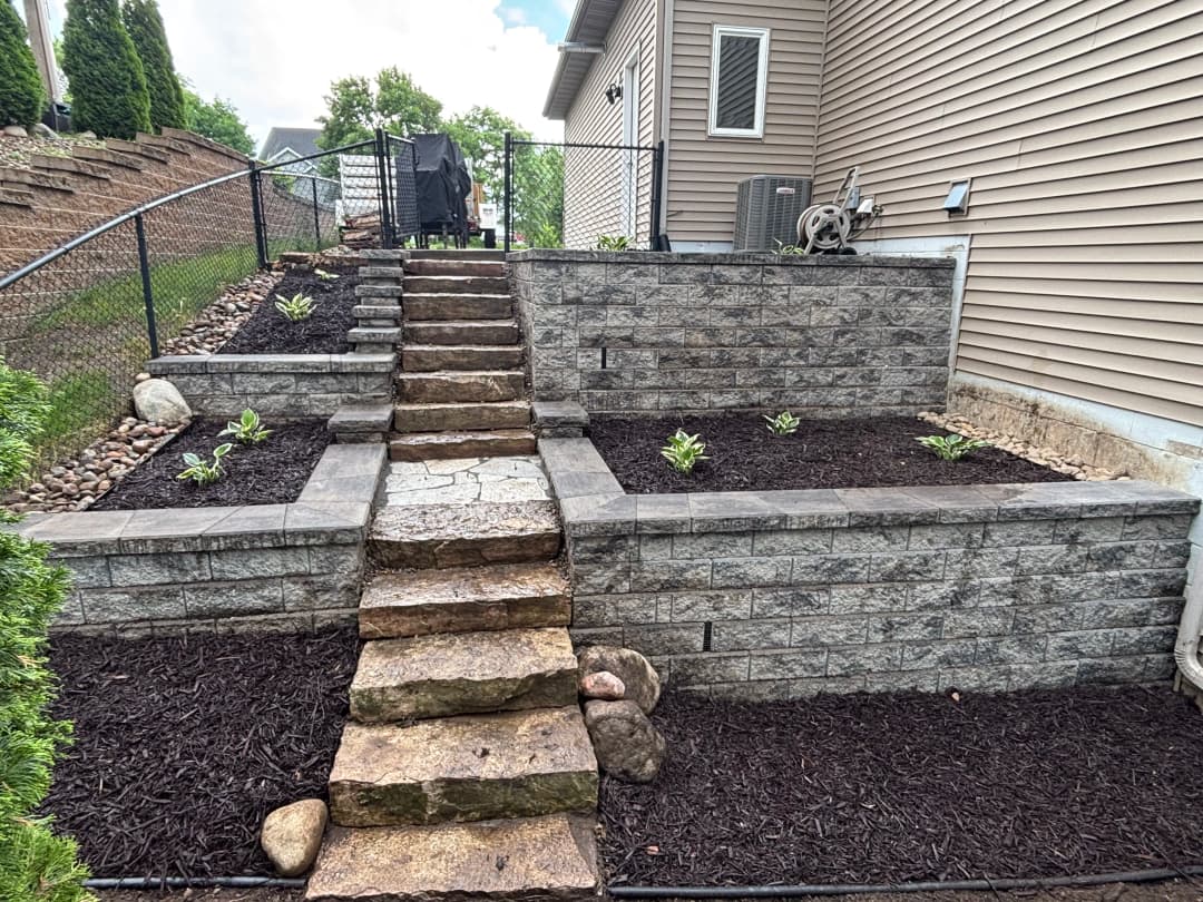 Stone steps leading to tiered garden beds with mulch and plants next to a house.