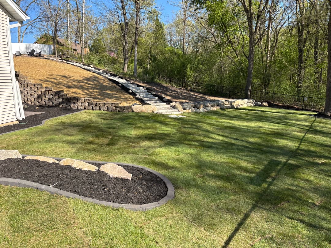 Lush green lawn with stone pathway and landscaped hill, surrounded by trees in springtime.