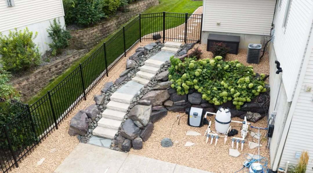 landscaped backyard with stone steps, plants, and pool equipment beside a fence