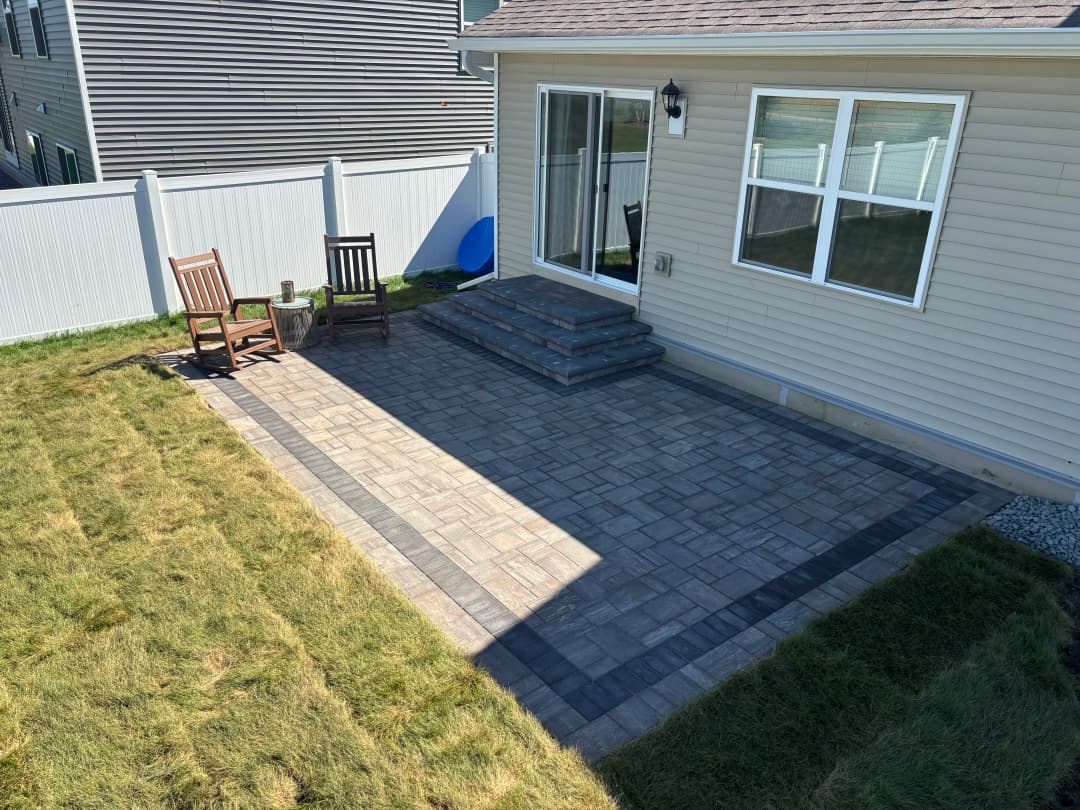 Outdoor patio with stone pavers, chairs, and steps next to a house and lawn.