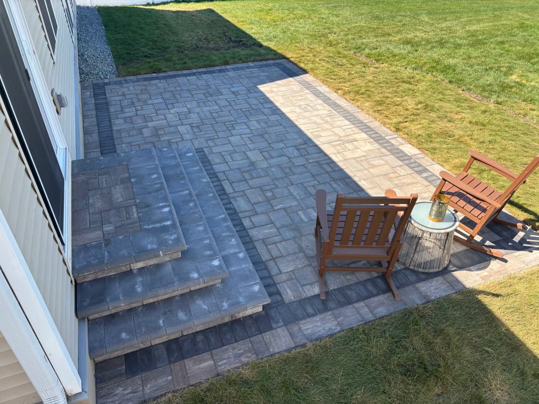 Patio with stone pavers and wooden chairs, surrounded by green grass and sunlight.