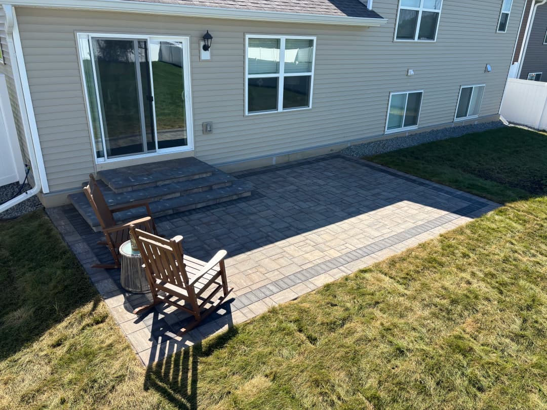 Backyard patio with stone pavers, wooden chairs, and grass area beside a house.